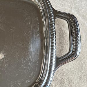 May include: Silver-toned rectangular serving tray with ornate embossed floral and scrollwork design. The tray has integrated handles and a decorative, ridged border. A classic piece for serving food and drinks.
