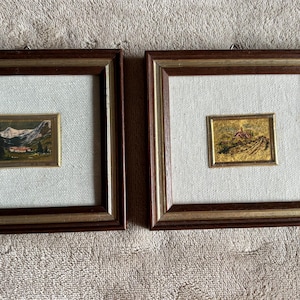 May include: Two square wall frames with dark brown and gold borders, each containing a light-colored fabric mat and a small, rectangular landscape painting. The left painting depicts a mountain scene, while the right shows a rural landscape.