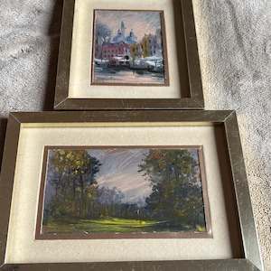 May include: Two framed paintings with gold-toned metallic frames and off-white matting. The top square painting depicts a cityscape with domes and buildings. The bottom rectangular painting shows a landscape with trees and a grassy field.