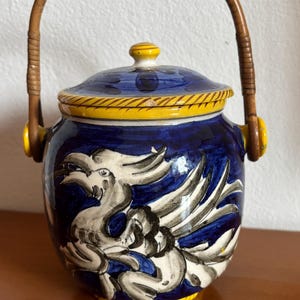 May include: A blue ceramic jar with a yellow rim and a dragon design. It has a lid and a woven handle. The lid has a yellow knob. The jar is decorated with white and black details.