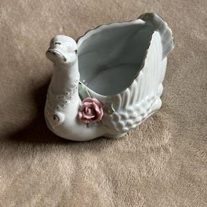 Pode incluir: Prato de cerâmica branca em forma de cisne com uma rosa rosa e detalhes dourados. O pescoço do cisne é curvo e o prato tem a forma do corpo de um cisne.
