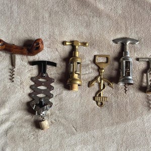 May include: A selection of six corkscrews with diverse designs. The corkscrews are made from materials such as wood, brass, and metal. They are displayed on a textured, light brown surface. Each corkscrew has a unique style.
