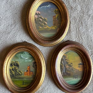May include: Three oval-shaped wall art pieces featuring hand-painted landscapes in gold-toned frames. Each painting depicts a scene with a structure, trees, and water. The frames have a distressed finish with visible red and gold paint layers.