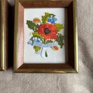 Op de afbeelding: Een rechthoekig wandobject met een bloemenborduurwerk op een witte stoffen achtergrond. Het ontwerp bevat een grote rode klaproos, groene bladeren en kleine blauwe bloemen, samengebonden met een lichtblauw lint. De lijst is gemaakt van donker hout met een dunne gouden metalen rand.