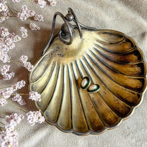 May include: A vintage brass shell-shaped dish with a handle. The dish is decorated with a ribbed pattern and has two small, oval-shaped objects inside.