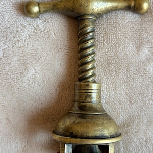 Op de afbeelding: Een messing kurkentrekker met een T-vormig handvat met afgeronde uiteinden, een centraal verticaal schroefmechanisme en een klokvormige behuizing. Het metalen oppervlak vertoont een verweerde, verouderde patina met zichtbare gebruikssporen.