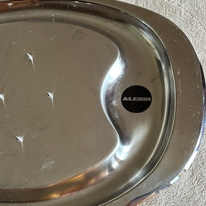 May include: A stainless steel serving tray with a polished finish. The oval-shaped tray has a slightly raised edge and the brand name "ALESSI" is visible on a black sticker. The tray is on a textured surface.
