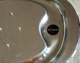 Può includere: Un vassoio da portata in acciaio inossidabile con finitura lucida. Il vassoio di forma ovale ha un bordo leggermente rialzato e il marchio "ALESSI" è visibile su un adesivo nero. Il vassoio è su una superficie strutturata.