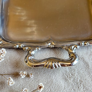 May include: A rectangular silver-toned metal serving tray featuring a smooth, reflective surface and scalloped edges. The tray includes a decorative, ornate handle with a shell-like motif at the center, providing a polished and metallic finish.