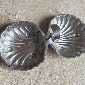 May include: A silver-coloured, hinged dish with a seashell design. The dish is made of metal and has a ring connecting the two halves. The dish is resting on a textured, light brown surface.