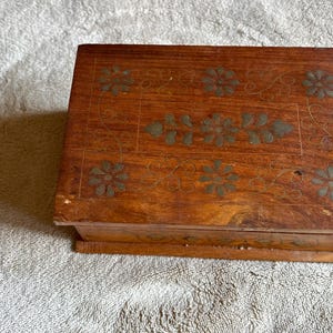 May include: A rectangular wooden box with a medium brown finish, featuring inlaid brass floral motifs on the lid. The design includes six individual flower shapes surrounding a central leaf and flower pattern, all connected by thin, etched lines.
