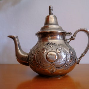 May include: A silver-toned metal teapot featuring an ornate embossed floral and scrollwork pattern on its rounded body. It has a curved spout, a hinged lid with a finial top, and a single handle. The surface has a polished, reflective finish.