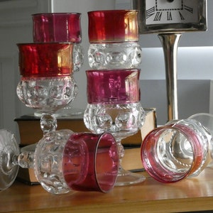 May include: Six ruby red glass goblets with a clear glass base and a textured pattern. The goblets are arranged on a wooden surface.