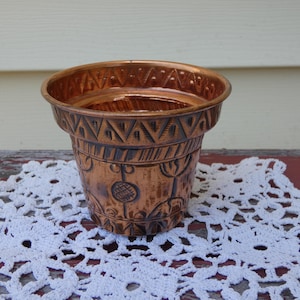 May include: A copper-colored, vintage-style planter with etched geometric and floral designs. The small, round pot has a wide rim and a slightly tapered body. The planter is sitting on a white lace doily.