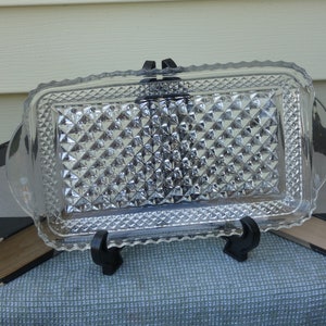 May include: A clear glass serving tray with a diamond pattern. The tray has a scalloped edge and two handles. The tray is sitting on a black stand.