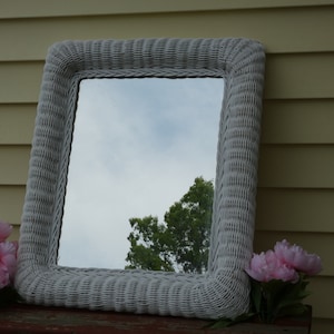May include: White wicker framed mirror with a rectangular shape. The mirror reflects a view of a tree and a cloudy sky.