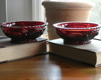 May include: Two red glass bowls with a decorative pattern sit on top of two books. The bowls are made of ruby red glass and have a scalloped edge.