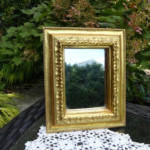 May include: A rectangular gold-colored mirror with an ornate, detailed frame. The frame has a textured surface with decorative patterns. The mirror reflects a view of trees and a building. The mirror is placed on a white lace doily.