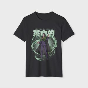 Op de afbeelding: Zwart t-shirt met een afbeelding van een personage in een paars pak en groene cape, omringd door groene energie wervelingen. De tekst "MONKEY D. DRAGON" en Japanse karakters staan boven het personage.