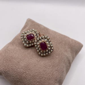 May include: A pair of gold earrings featuring a ruby gemstone set within a halo of pearls.