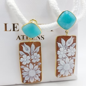 Puede incluir: Pendientes dorados con tapas cuadradas turquesas y colgantes rectangulares marrones. Los colgantes presentan tallas florales blancas. Los pendientes se muestran sobre un fondo blanco.