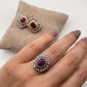 Peut inclure: Une bague avec un gros rubis ovale entouré de petites perles blanches et de diamants. La bague est sertie sur une bande en or.