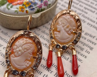 May include: Gold tone earrings with a cameo design featuring a woman's profile. The earrings have a dangling design with three red coral beads on each earring.