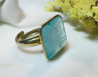 May include: A gold-toned adjustable ring featuring a large, square-cut, light blue gemstone. The gemstone is set in a gold bezel, and the ring is displayed on a white surface with yellow flowers in the background.