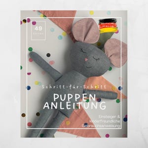 May include: A gray plush mouse doll with pink ears and a pink heart nose, lying on a white background with colorful confetti. The image includes text that reads "PUPPEN ANLEITUNG" and "DEUTSCHE VERSION". The doll is part of a sewing pattern.