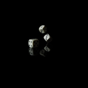 Może przedstawiać: Trzy białe kostki do gry z czarnymi kropkami zawieszone w powietrzu na czarnym tle. Jedna kostka spoczywa na odblaskowej powierzchni, a pozostałe dwie są w ruchu, tworząc dynamiczną kompozycję. Kostki do gry są używane do gier.