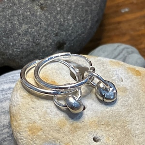 May include: A pair of silver hoop earrings with small silver charms hanging from them. The earrings are on a light brown rock.