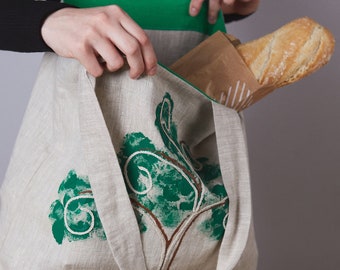 Może przedstawiać: Naturalna lniana torba z zielono-brązowym wzorem drzewa. Torba ma zielone wnętrze i mieści bagietkę w brązowej papierowej torbie. Torba ma długie paski.