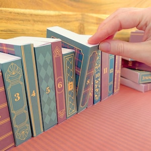 Op de afbeelding: Een rij kleine, rechthoekige doosjes ontworpen als miniatuurboeken, elk met een uniek patroon op de rug in tinten groen, blauw en bordeauxrood, voorzien van goudkleurige lijnen en de cijfers 3 tot en met 12 op de voorzijde gedrukt.