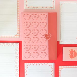 May include: A collection of pink stationery items, including notepads and an envelope, arranged on a red surface. The central notepad features heart-shaped candies with phrases like "Love You" and "Kiss Me." Other notepads have lined pages and decorative borders.