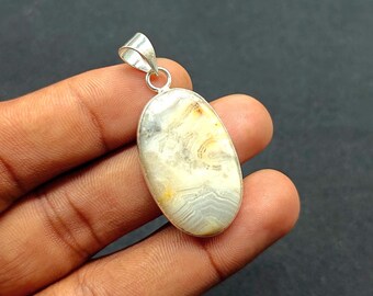 May include: Oval-shaped pendant with a silver bail. The pendant features a polished stone with a mix of colors, including white, gray, and yellow. The stone has a unique, natural pattern. The pendant is held in a hand.