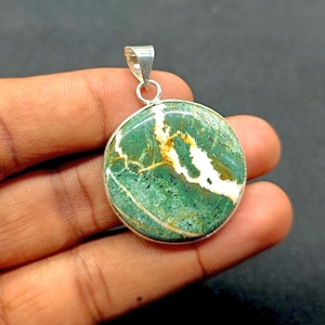 Op de afbeelding: Ronde hanger met een groene steen, met witte en gele adering. De steen is gezet in een zilverkleurig metalen frame met een lus voor een ketting. De hanger wordt in een hand gehouden tegen een donkere achtergrond.