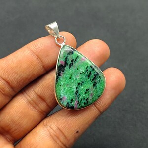 Puede incluir: Un colgante en forma de lágrima con una fianza plateada. El colgante presenta una piedra verde vibrante con inclusiones negras y rojas. La piedra está engastada en un bisel plateado.