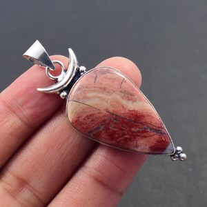 Op de afbeelding: Een druppelvormige hanger met een rood en crèmekleurige steen, met donkerrode adering. De steen is gezet in een zilverkleurig metalen frame met een halve maan ontwerp aan de bovenkant en kleine decoratieve accenten.
