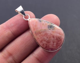 May include: A teardrop-shaped pendant with a pink gemstone, set in a silver metal frame. The gemstone has a textured appearance with darker spots. The pendant has a silver bail for attaching to a chain.
