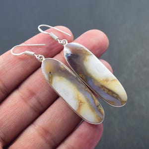 May include: A pair of silver-toned dangle earrings. Each earring features an elongated, oval-shaped stone with a marbled pattern of white, brown, and lavender hues. The earrings are held by a person's hand against a dark background.