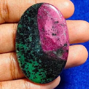 Peut inclure: Une pierre précieuse ovale polie. La pierre présente une combinaison de couleurs frappante de rouge profond, de noir et de vert. La pierre est tenue dans une main sur un fond bleu, soulignant sa beauté naturelle.