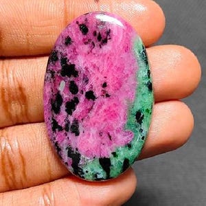 Puede incluir: Gema ovalada con un centro rosa vibrante, moteado con inclusiones negras y bordeado por un borde verde. La piedra pulida se sostiene en una mano, mostrando sus colores y patrones naturales. Ideal para la creación de joyas.