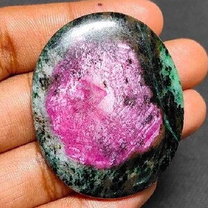 May include: An oval gemstone featuring a bright pink core surrounded by a dark green and black border. The polished stone is held in a hand, highlighting its natural beauty and unique colour pattern. A striking piece for jewellery.
