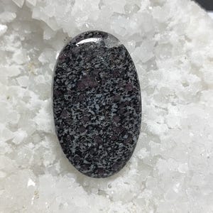 May include: An oval gemstone with a speckled pattern of black, grey, and burgundy. The polished surface reflects light, highlighting the stone's natural texture. The gemstone is set against a backdrop of white, crystalline material.