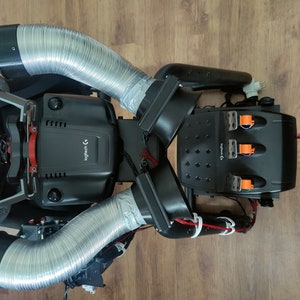 May include: Overhead view of a black racing simulator setup with silver flexible tubing and the Logitech logo. The setup includes a steering wheel, pedals, and other controls, all mounted on a black frame.