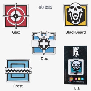 May include: Set of 6 enamel pins featuring characters from the video game Rainbow Six Siege. The pins are square shaped with a black outline and feature the characters Glaz, Blackbeard, Doc, Frost, Ela, and a Rainbow Six Siege logo.
