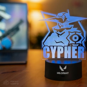 Op de afbeelding: Een blauwe en witte 3D-geprinte lamp met een personage uit de videogame Valorant. Het personage is Cypher, en de lamp heeft het Valorant-logo op de basis.
