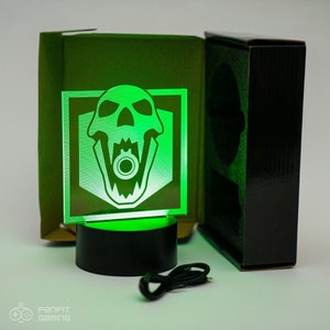 May include: A green light-up skull with a gun in its mouth, in a black base, with a USB cord. The skull is in a white square with a black border. The light is on and the skull is glowing green.