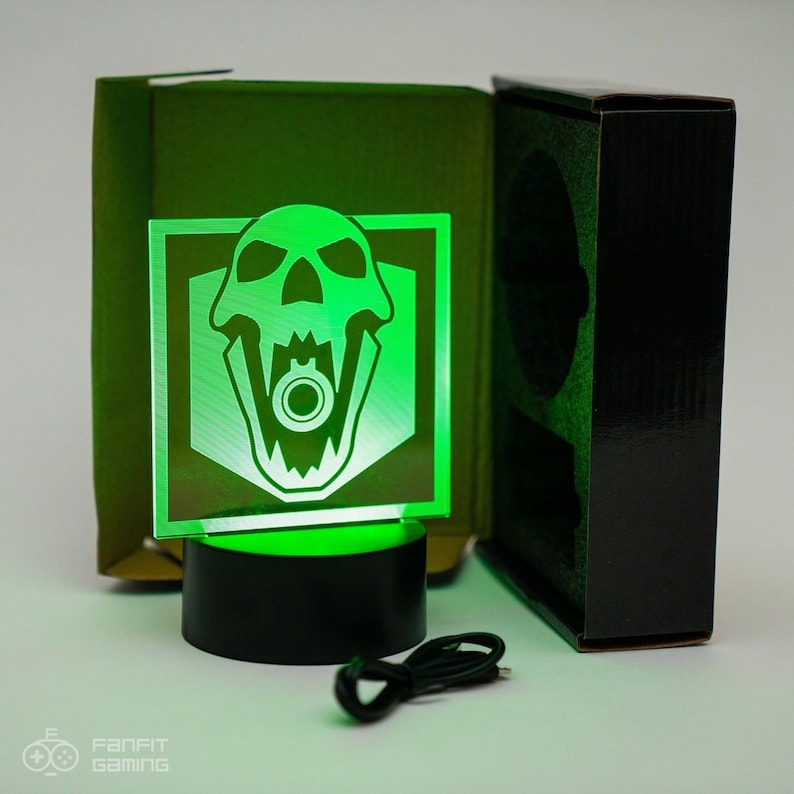 May include: A green light-up skull with a gun in its mouth, in a black base, with a USB cord. The skull is in a white square with a black border. The light is on and the skull is glowing green.