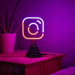 May include: A neon sign shaped like the Instagram logo. The sign is lit up in pink and white and is sitting on a black base. The sign is on a wooden table.